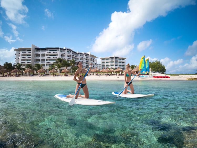 Two women on paddle boards in water, resort building in the background