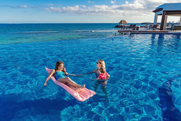 a girl on inflatable pad in pool, another woman holding her baby, ocean view in background